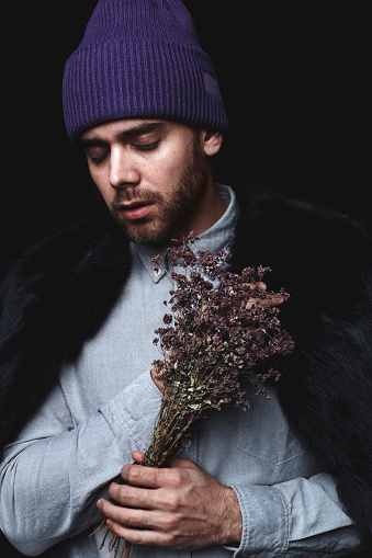 Portrait of young handsome bearded man in fur coat and violet hat holding bouquet of flowers with his eyes closed