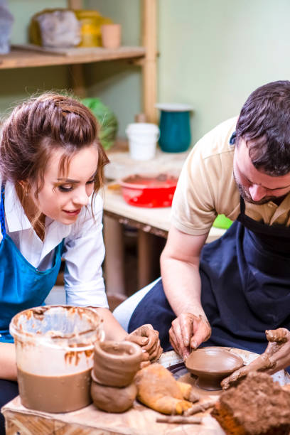 istock Male Potter Tutor Working with Female Apprentice with Clay Lump on Potter's Wheel in Workshop.Vertical Orientation 1272731014