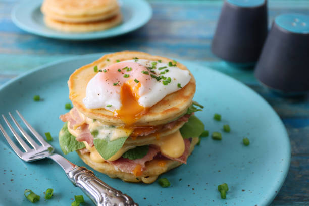istock Close-up image of turquoise blue plate, metal fork,mini pancakes stack, florentine eggs benedict recipe, layers of pancake, bacon, spinach leaves, hollandaise sauce, poached egg, salt cellar, pepper pot, blue wood background, focus on foreground 1272738537