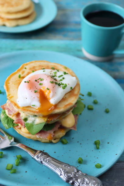 istock Image of turquoise blue plate, metal fork and stack of mini pancakes, florentine eggs benedict recipe, layers of pancake, bacon, spinach leaves, hollandaise sauce, poached egg oozing egg yolk, mug black coffee, blue wood background, focus on foreground 1272738539