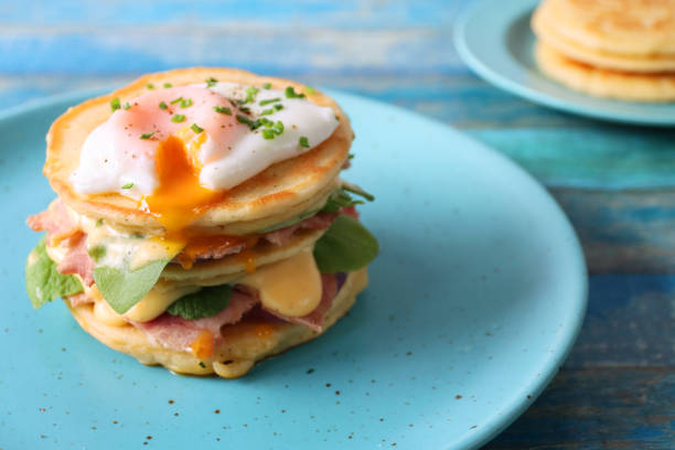 istock Close-up image of turquoise blue plate with stack of mini pancakes, florentine eggs benedict recipe, layers of pancake, bacon, spinach leaves, hollandaise sauce and poached egg with oozing egg yolk, blue wood background, focus on foreground 1272738559