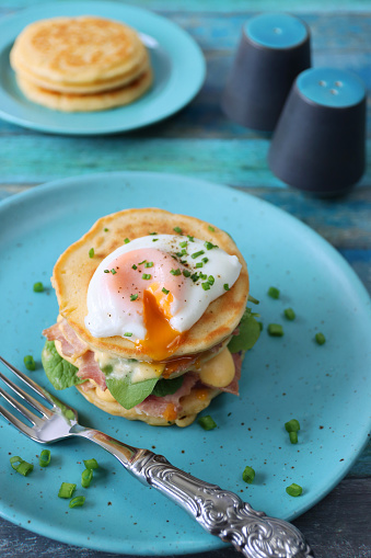 Stock photo showing a close-up view of a turquoise plate containing a stack of mini pancakes, served florentine eggs benedict style against a blue wood background. Layers of pancake, bacon, spinach leaves, hollandaise sauce and poached egg beside a metal fork, salt cellar and pepper pot.