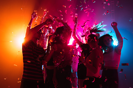 Rear view of large group of people enjoying a concert performance. There are many hands applauding and taping the show. Multi colored lasers and spot lights firing from the stage.\nSilhouettes have been significantly liquified.