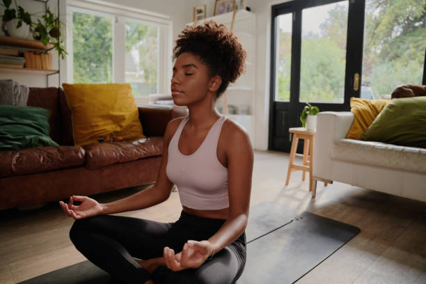 Young black woman doing yoga at home in the lotus position Young woman sitting on yoga mat meditating meditating photos stock pictures, royalty-free photos & images