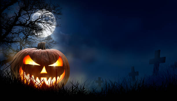 A spooky halloween pumpkin, Jack O Lantern, with an evil face and eyes. A spooky halloween pumpkin, Jack O Lantern, with an evil face and eyes on the grass in a graveyard with a misty night sky background with a full moon. halloween pumpkin jack o lantern horror stock pictures, royalty-free photos & images