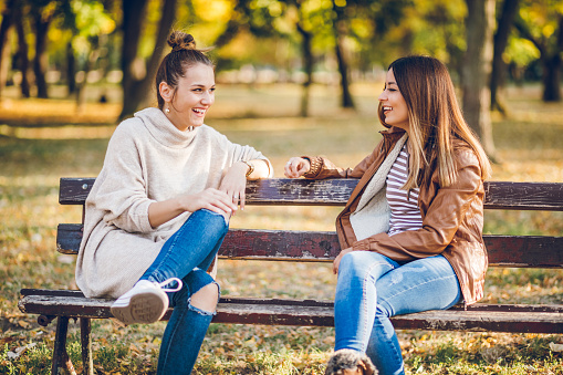 Two young caucasian beautiful women sitting on park bench and talking. Autumn time.
