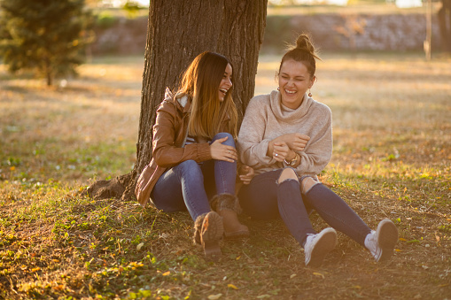 Two young beautiful caucasian women sitting under a tree, talking and laughing. Autumn time.