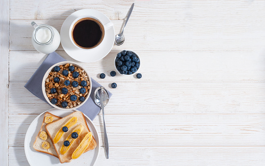 Breakfast with granola and blueberry, coffee, milk with orange juice and baked toasts on the white wooden table flat lay with copy space. Healthy tasty food at the morning with oat-flakes