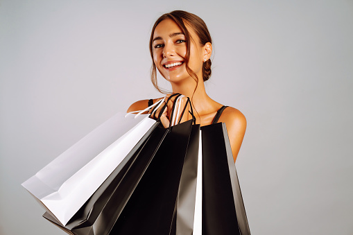 Elegant brunette woman in black dresse holding shopping bags. Young woman with black and white bags posing on grey background. Purchases, black friday, discounts, sale concept.