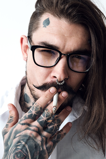 Headshot of smoking tattooed man. Portrait of stylish middle-aged hipster with long hair, tattoos, moustache and pierced nose smoking a cigarette and looking at camera impudently