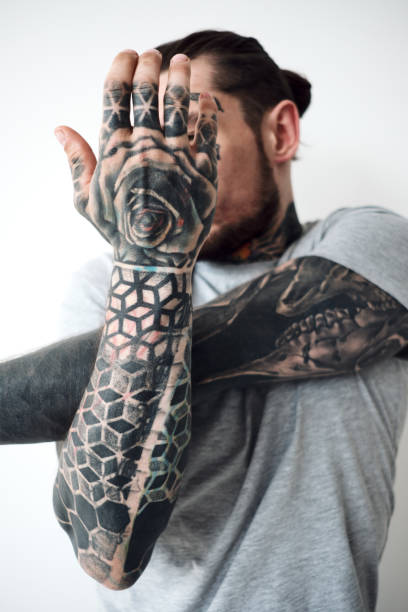 Conceptual shot of tattooed man. Low angle view of bearded man with tattooed arms standing in unnatural pose and hiding his face against white wall background