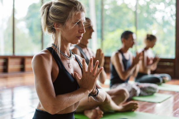 Woman clasping hands during yoga lesson Adult female clasping hands and looking away while meditating during group yoga lesson in light studio yoga class stock pictures, royalty-free photos & images