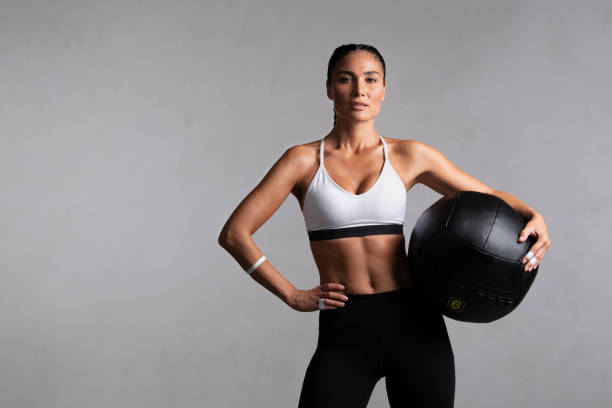 Determined fitness woman on grey background Portrait of beautiful mid adult woman looking at camera while holding heavy medicine ball isolated on grey background. Proud and fit woman standing on gray wall ready for gym exercise while looking at camera. Strong mixed race girl holding sports ball and relaxing after cross training workout with copy space. weight training stock pictures, royalty-free photos & images