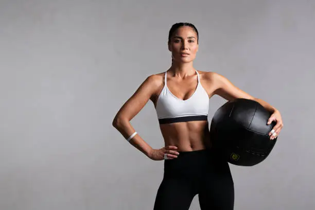 Portrait of beautiful mid adult woman looking at camera while holding heavy medicine ball isolated on grey background. Proud and fit woman standing on gray wall ready for gym exercise while looking at camera. Strong mixed race girl holding sports ball and relaxing after cross training workout with copy space.