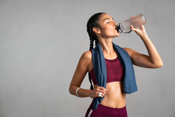 Fit woman drinking from water bottle Fitness thirsty girl drinking water standing on gray background with copy space. Portrait of sweaty latin woman with towel around neck take a break after intense workout. Mid adult fit woman taking a break and drink from water bottle after gym workout isolated on grey wall. energy drink photos stock pictures, royalty-free photos & images