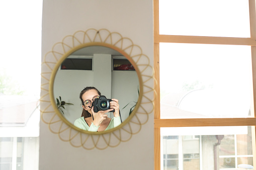 Young woman with camera in hands standing in front of the mirror and taking photo of herself