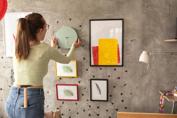 istock Young woman placing watch on wall while decorating living room 1272783064
