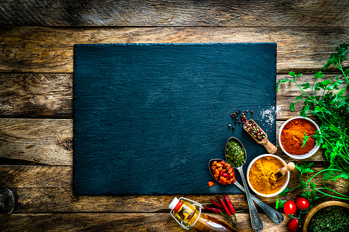 Food backgrounds: overhead view of multi colored spices, herbs and vegetables arranged at the bottom right corner of a rustic wooden table. An empty slate board is at the center leaving useful copy space for text and/or logo. Spices and herbs included in the composition are dried parsley, lime, chili pepper, peppercorns, nutmeg, olive oil, turmeric, ground paprika, salt, tomatoes, among others. High resolution 16:9 format 35,5Mp studio digital capture taken with SONY A7rII and Zeiss Batis 40mm F2.0 CF lens
