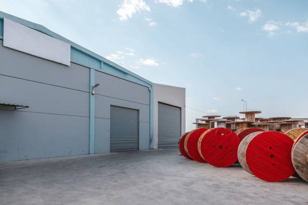 Large wooden bobbins with cable wire against factory warehouse outdoor Large and red colour wooden cable spools stands in front of large electronic cable factory warehouse outside on sunny day wooden spool stock pictures, royalty-free photos & images
