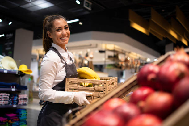 je travaille à l’épicerie. ouvrier de supermarché fournissant le département de fruit avec la nourriture. ouvrier féminin retenant la caisse avec des fruits. - commerçant photos et images de collection