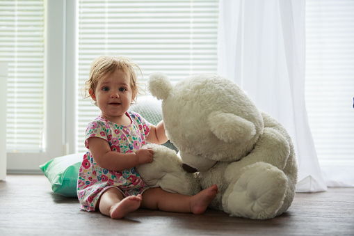 Full length shot of adorable baby girl sitting on the nursery floor and looking at camera with excitement while playing with her big teddy bear toy.