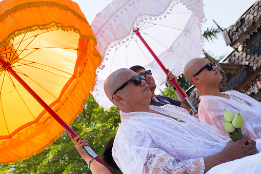 Bald headed thai men with sunglasses at ceremony to be monk are sitting under parasols on a pick-up. People are bringing and accompanying men to temple to be monks once in life.