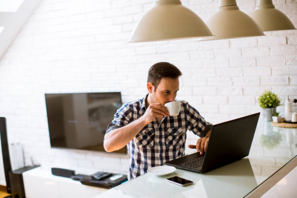 istock Young man sitting on kitchen desk at home and using laptop and drinking coffee 1272791432