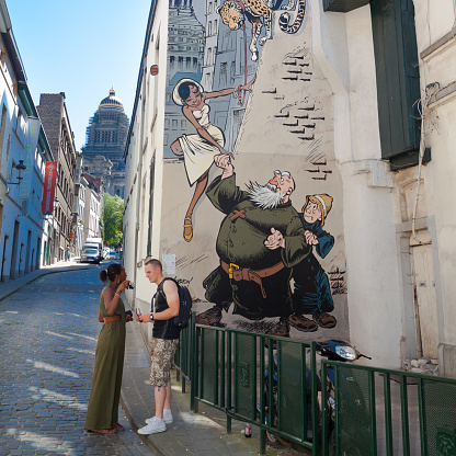 Caucasian man and black woman are standing in street Rue des Capucins in Brussels. Street is small street in district Marolles. People are talking. Woman is holding a camera in hand and man is looking at display. In background is cartoon mural by Odilon Verjus with brother and monk Verron & Yann together with a black woman and a leopard. Painting is part of official tourist and comic / cartoon route of Brussels