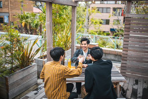 Smiling friends in early 20s wearing casual clothing and sitting at picnic table on Tokyo building terrace holding up glasses for toast.