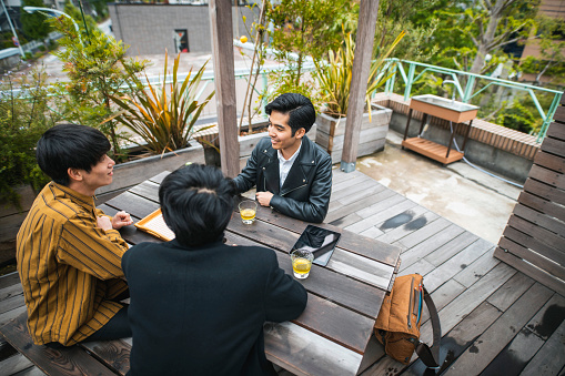 Elevated view of smiling young Japanese men in casual clothing sitting and talking outside on building terrace.