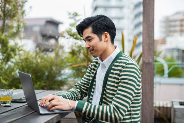 istock Young Japanese Man Typing on Laptop While Sitting Outdoors 1272799768