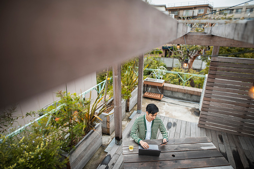 Personal perspective of male writer in early 20s sitting at outdoor picnic table and pausing to check smart phone while typing on laptop.