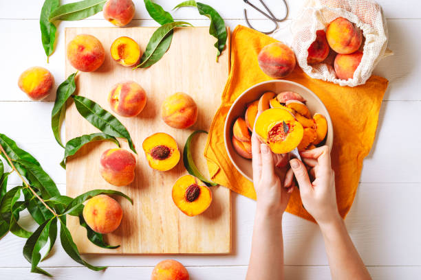 Female hands cutting fresh sweet peaches. Peaches whole fruits leaves, half peach, peach slices on white wooden kitchen table. Recipe making peach jam, cooking peach dessert on cutting board.Flat lay. manos femeninas cortando melocotones dulces frescos. melocotones hojas enteras de frutas, medio melocotón, rodajas de melocotón en la mesa de cocina de madera blanca. receta haciendo mermelada de melocotón, cocinar el postre de melocotón en tabla de co