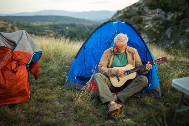 istock Senior man sitting in front of tent and playing guitar while camping 1272821111