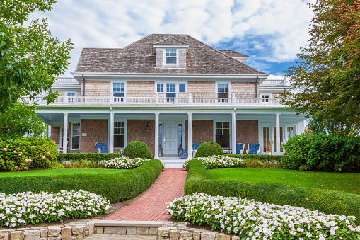 Newly Renovated New England Mansion with Grey Shingle Exterior, Grey Roof and Landscaped Front Yard with Red Footpath, green bushes and white flowers. Blue chairs are on the porch. The house is in Chatham, Cape Cod, Massachusetts. Canon EF 24-105mm/4L IS USM Lens.