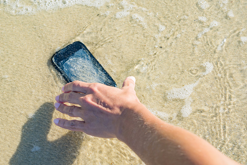 Woman pulls out a dropped smartphone from the sea. Finding lost things, repair of drowned mobile phones, concept image.