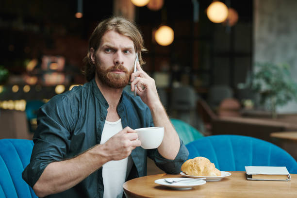 istock Busy young bearded man sitting at table in modern cafe and drinking coffee while discussing project by phone 1272830921
