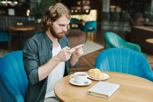 istock Serious handsome young man with beard sitting at table and calling colleague by phone while waiting for him in cafe 1272831109