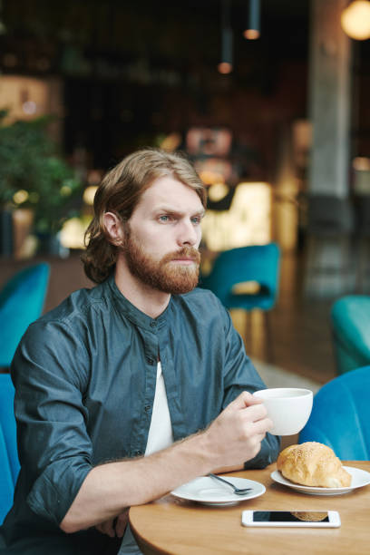 istock Serious handsome guy with beard sitting at table in cafe and drinking coffee while reading mail on smartphone 1272831245