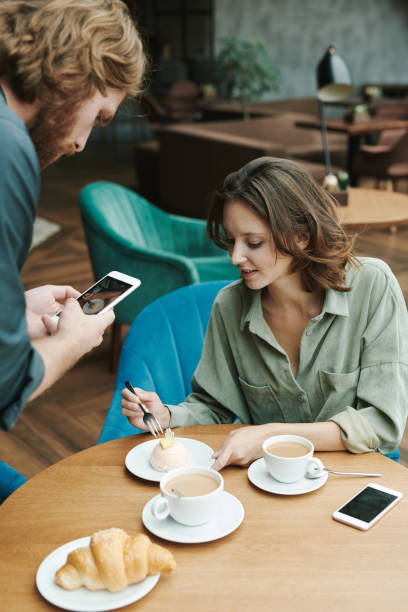 istock Busy young bearded man sitting at table in modern cafe and drinking coffee while discussing project by phone 1272833011