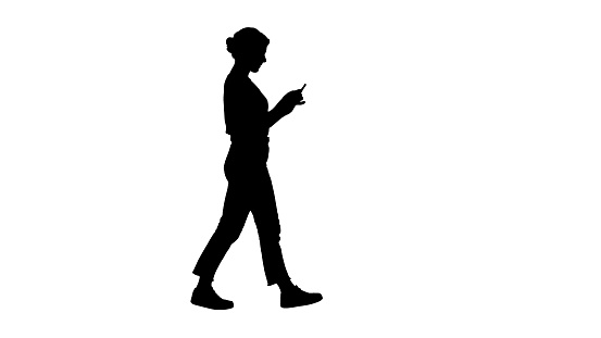 Full length shot. Side view. Silhouette Beautiful young woman walking and reading text message on her mobile phone. Professional shot in 4K resolution. 006. You can use it e.g. in your commercial video, business, presentation, broadcast