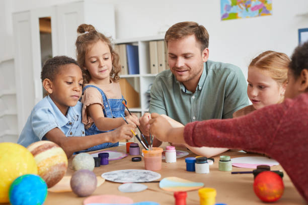 Kids Enjoying Paining in Art and Craft Class Portrait of multi-ethnic group of children holding brushes and painting planet model while enjoying art and craft lesson in school or development center, copy space preschool teacher stock pictures, royalty-free photos & images