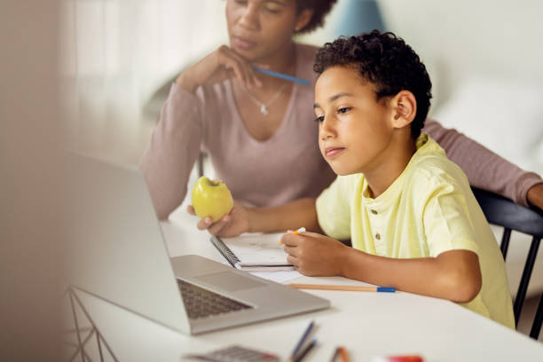 Little black boy using laptop while homeschooling with his mother. African American boy eating apple and writing notes while e-learning over laptop at home. food elementary student healthy eating schoolboy stock pictures, royalty-free photos & images