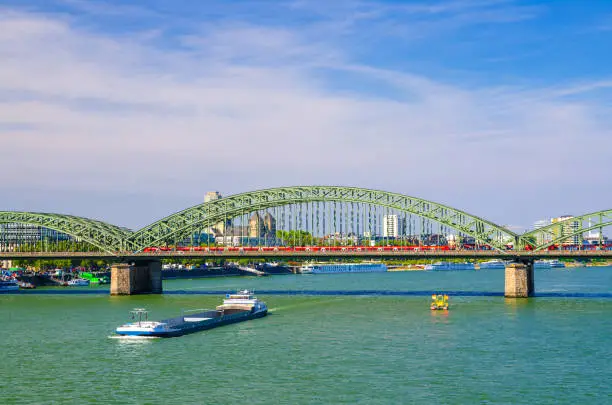 The Hohenzollern Bridge or Hohenzollernbrucke across Rhine river with cargo ships sailing on water, pedestrian and railway steel bridge, Cologne city centre, North Rhine-Westphalia, Germany