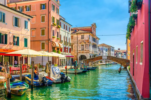 Photo of Chioggia cityscape with narrow water canal Vena, moored multicolored boats, street restaurant on embankment, old colorful buildings and brick bridge, blue sky in summer day, Veneto Region, Italy