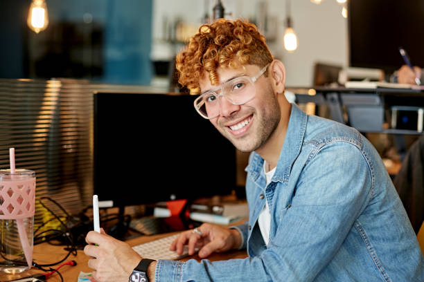 Portrait of Young Hispanic Freelancer in Coworking Office Man in late 20s with curly red hair, eyeglasses, and wearing casual clothing sitting at desktop computer and pausing from work to smile at camera. half shaved hairstyle stock pictures, royalty-free photos & images