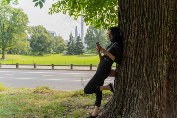 Teenage girl standing next to the tree and texting or checking social media with a smartphone in Central Park, Manhattan, New York. A 17-years-old teenage girl wearing protective mask in Central Park, Manhattan, NY black hair emo girl stock pictures, royalty-free photos & images