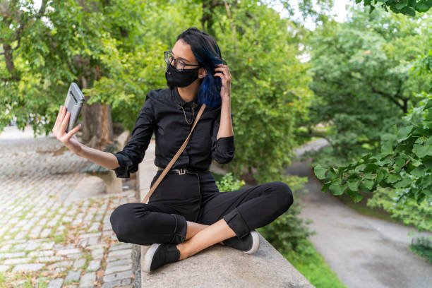 Teenage girl is sitting on the stone fence of Central Park, Manhattan, and taking selfies with a smartphone. A 17-years-old teenage girl wearing protective mask in Central Park, Manhattan, NY black hair emo girl stock pictures, royalty-free photos & images