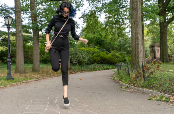A teenage girl playing hopscotch in Central Park, Manhattan, New York, deserted because of the COVID-19 pandemic. She is wearing a protective mask but holds in just on the neck because nobody is around. A teenage girl playing hopscotch in Central Park, Manhattan, New York black hair emo girl stock pictures, royalty-free photos & images