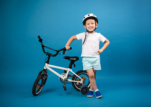 A child toddler with a child's bike and in protective helmet stands on a blue background and looks into the camera. Studio photo for articles on safety and kid sports and activity.